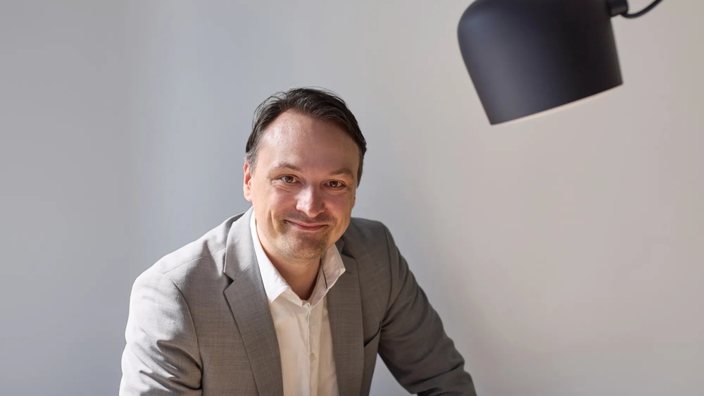 Ein Mann in grauem Anzug lächelt in die Kamera, vor einem hellen Hintergrund.