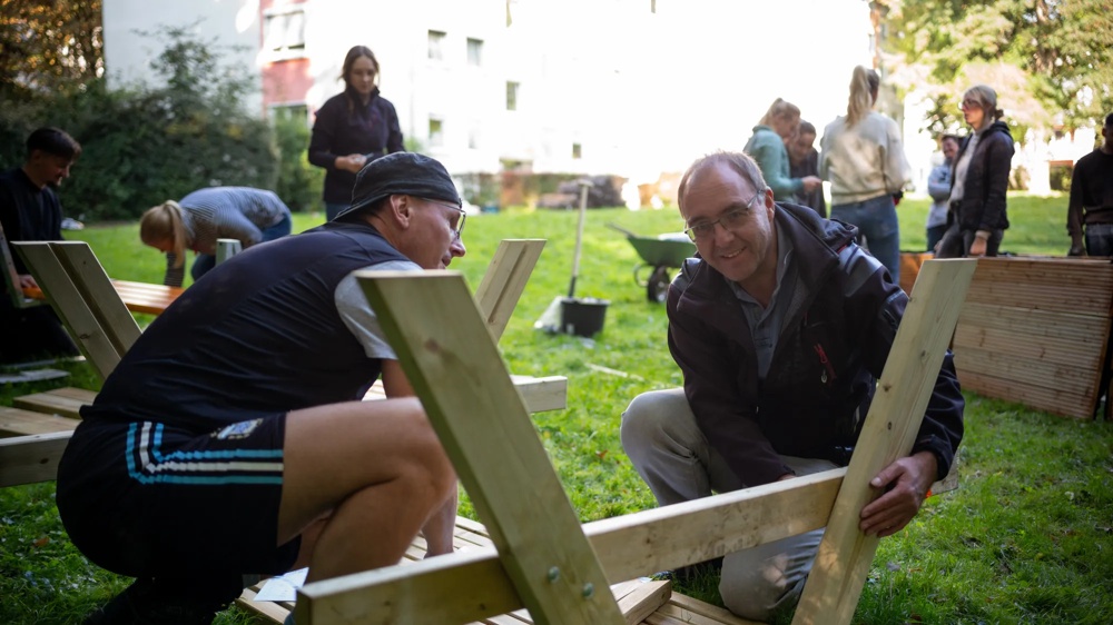 Two men assembling wooden furniture outdoors, surrounded by people on a green meadow.