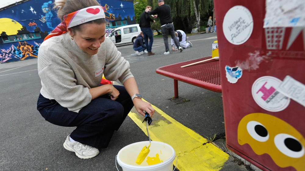 Eine Frau bemalt mit einer gelben Farbrolle eine Straße neben einer roten Bank. Im Hintergrund sind Graffiti zu sehen.