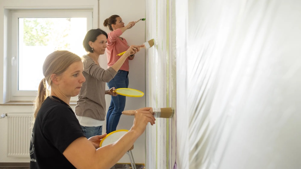 Three women painting a wall with brushes and yellow paint bowls.