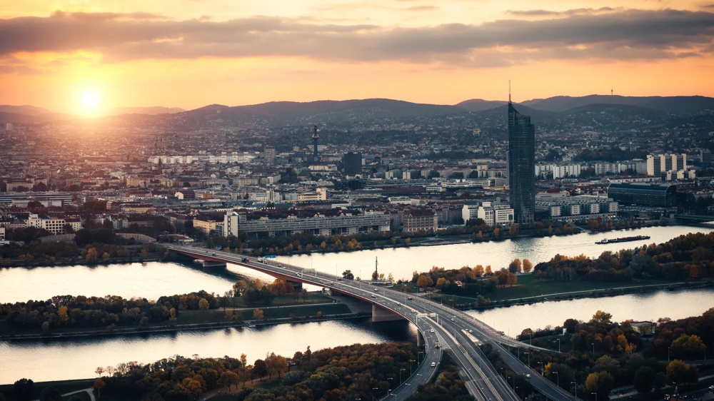 Sonnenuntergang über Stadt mit Fluss, Brücke und Hochhaus, umgeben von Hügeln.