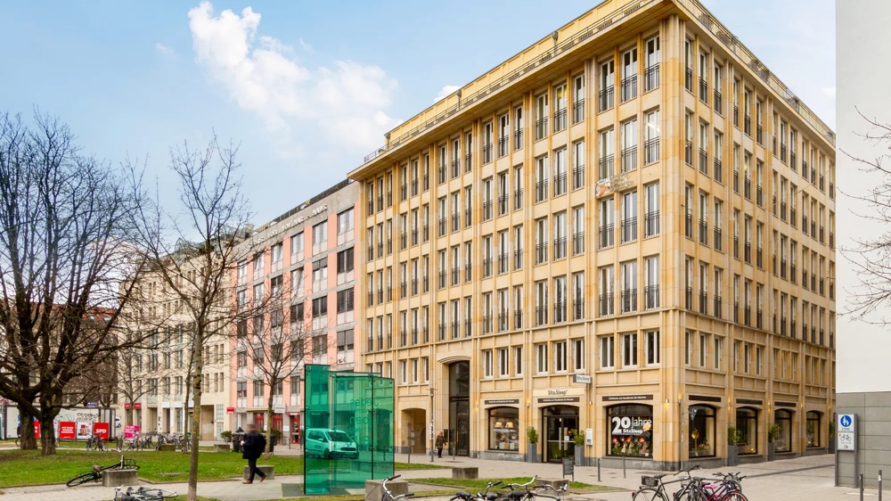 Ein mehrstöckiges, beiges Gebäude mit vielen Fenstern, umgeben von Bäumen und Fahrrädern, unter blauem Himmel.