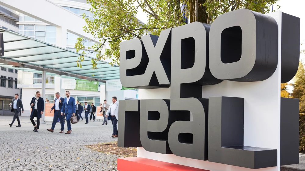 Großes "Expo Real"-Schild im Freien, Menschen in Anzügen gehen auf einem gepflasterten Weg vorbei.