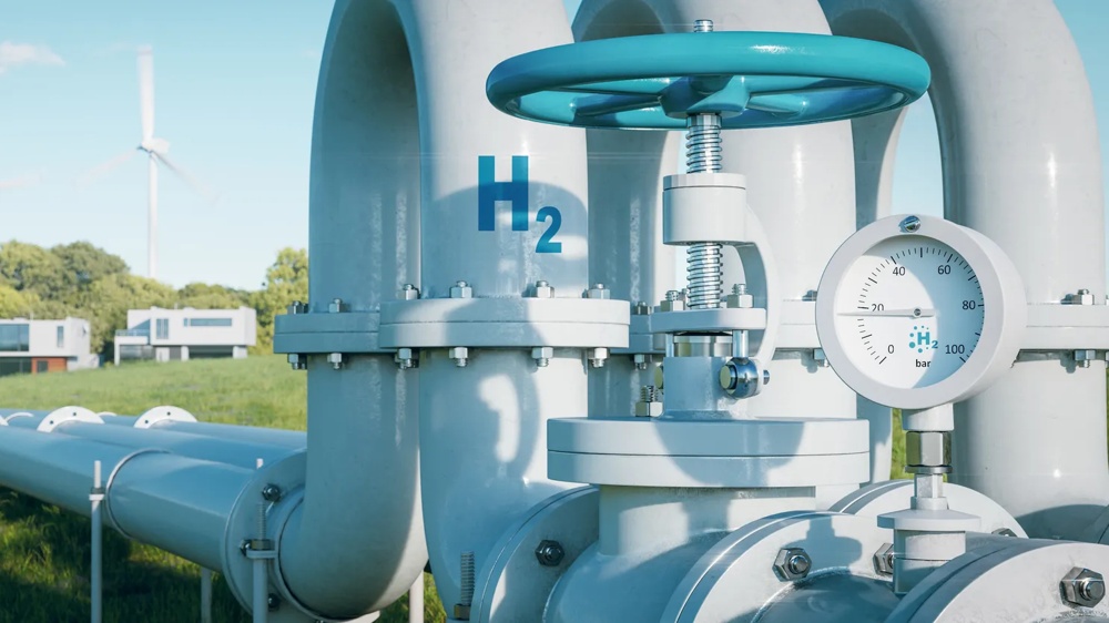 Große graue Wasserstoffleitungen mit Ventil und Manometer, Windräder und Gebäude im Hintergrund.