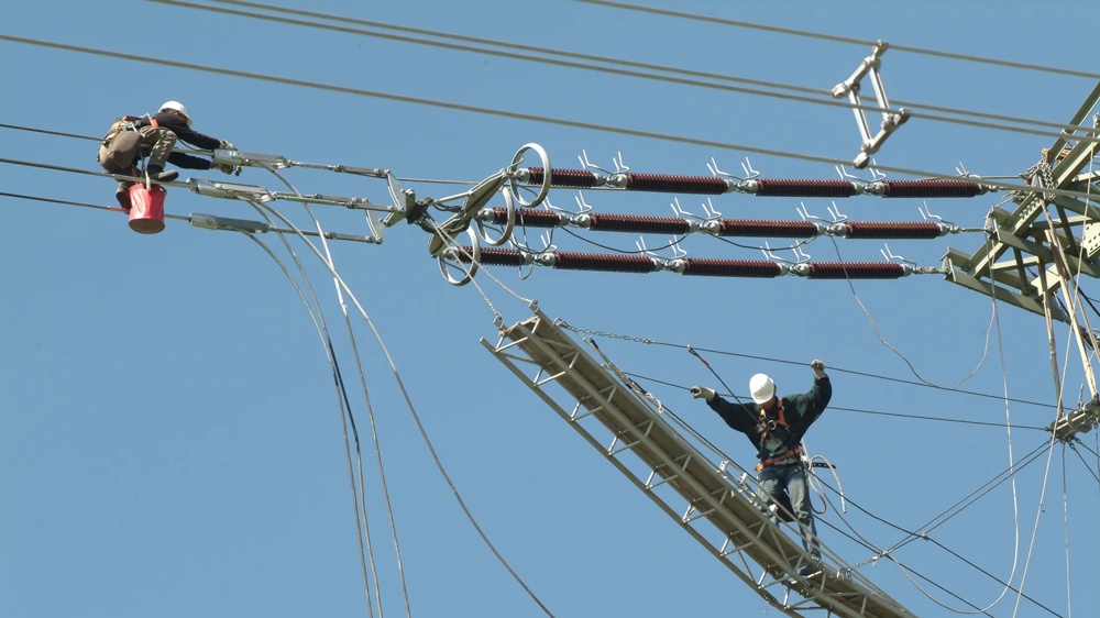 Zwei Arbeiter in Schutzkleidung reparieren Stromleitungen auf einem Hochspannungsmast vor blauem Himmel.