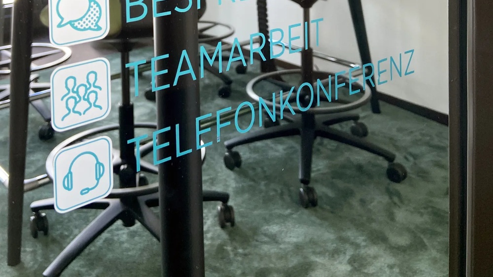 Text on glass wall with “Meeting” behind it, a conference table with chairs and a plant motif on the wall.