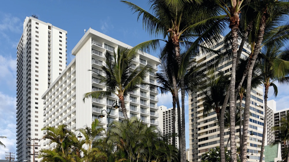Hohe weiße Gebäude mit Balkonen hinter Palmen vor blauem Himmel.