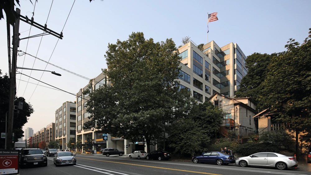 Mehrstöckiges Gebäude mit US-Flagge, Bäume und parkende Autos an der Straße bei klarem Himmel.