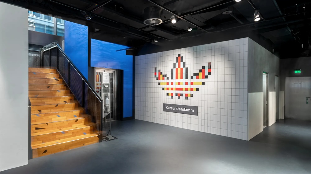 Holztreppe neben weißer Kachelwand mit pixelartigem Logo und "Kurfürstendamm"-Schild, moderner Innenraum.