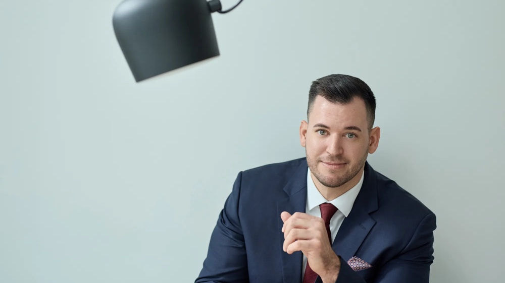 Ein Mann trägt einen schwarzen Anzug mit einer roten Krawatte und einem Einstecktuch. Er sitzt vor einer mintfarbenen Wand, neben ihm ragt eine schwarze Lampe ins Bild.