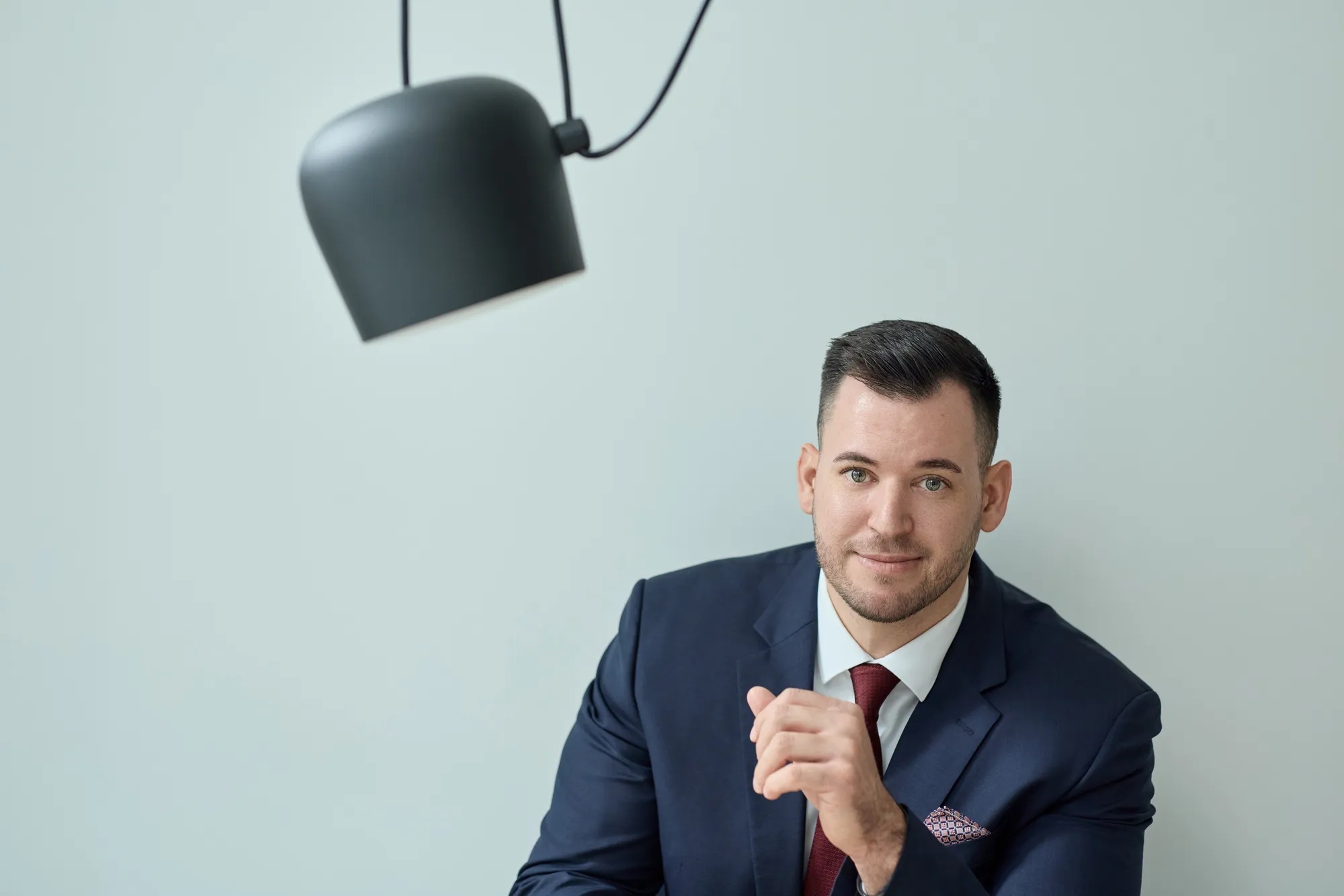 Ein Mann trägt einen schwarzen Anzug mit einer roten Krawatte und einem Einstecktuch. Er sitzt vor einer mintfarbenen Wand, neben ihm ragt eine schwarze Lampe ins Bild.