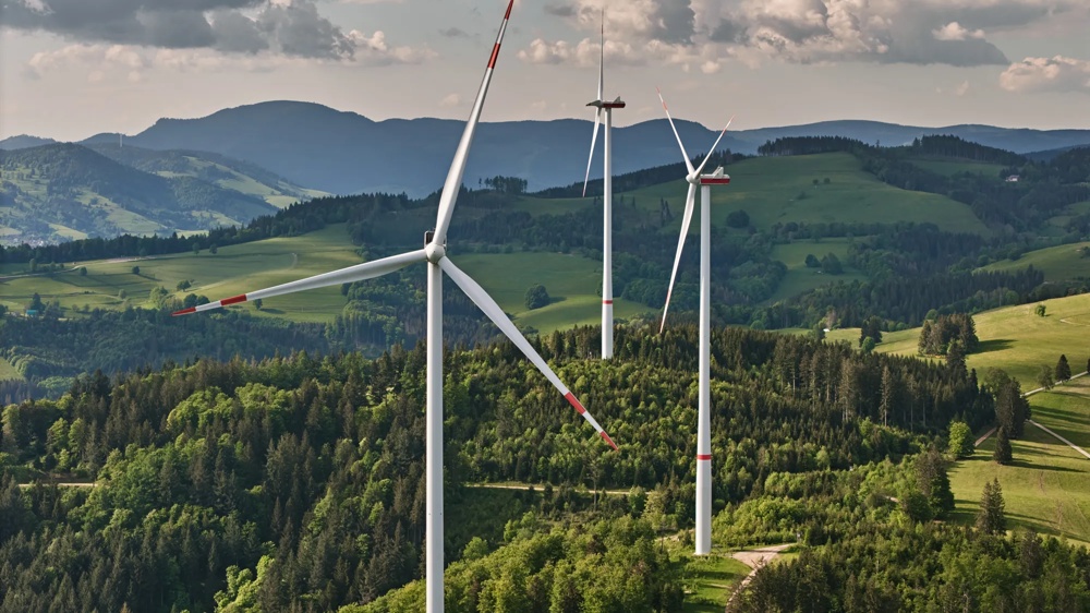 Drei Windräder auf einem bewaldeten Hügel unter bewölktem Himmel, umgeben von grünen Feldern und Bergen.