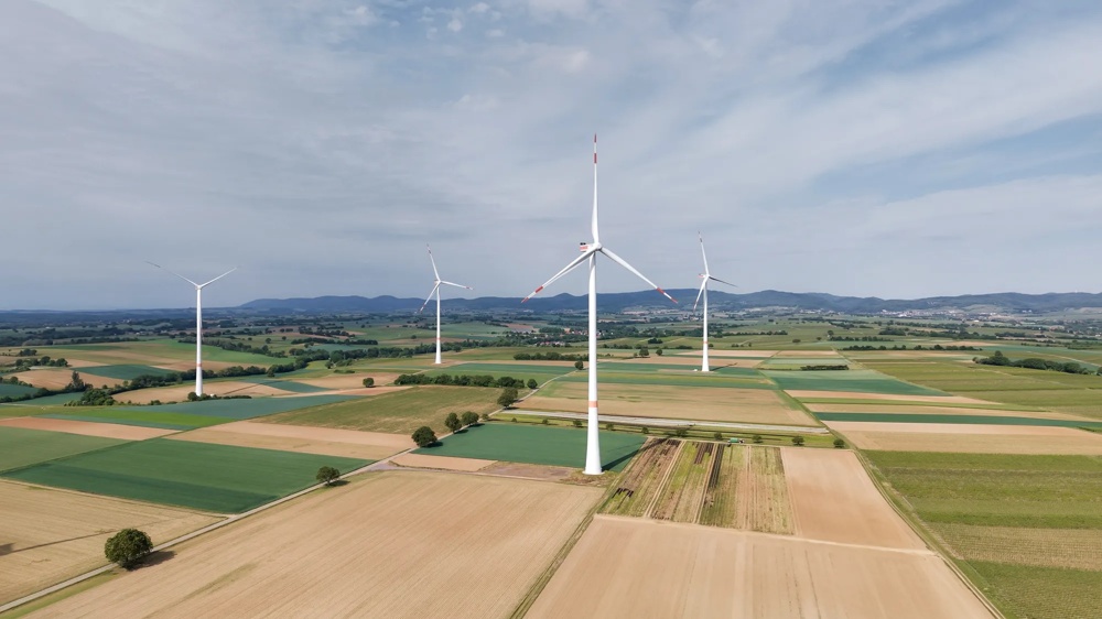 Windräder auf Feldern bei Sonnenuntergang, mit Hügeln im Hintergrund und dramatisch gefärbtem Himmel.