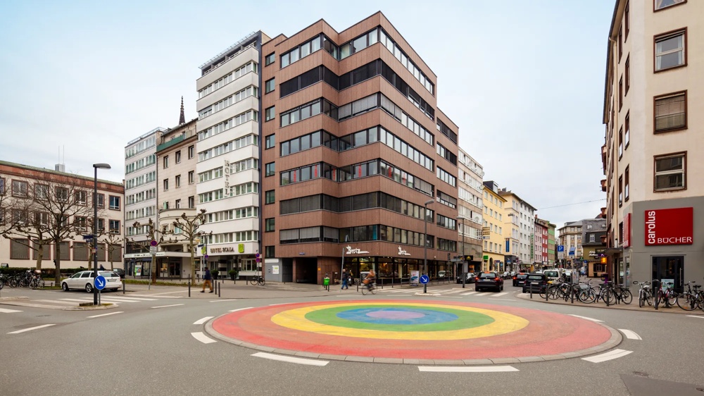 A rainbow-coloured roundabout surrounded by modern buildings and shops on a city street.