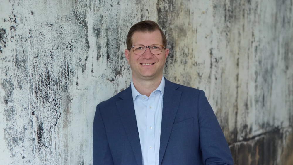 A man in a blue suit standing smiling in front of a weathered wall.