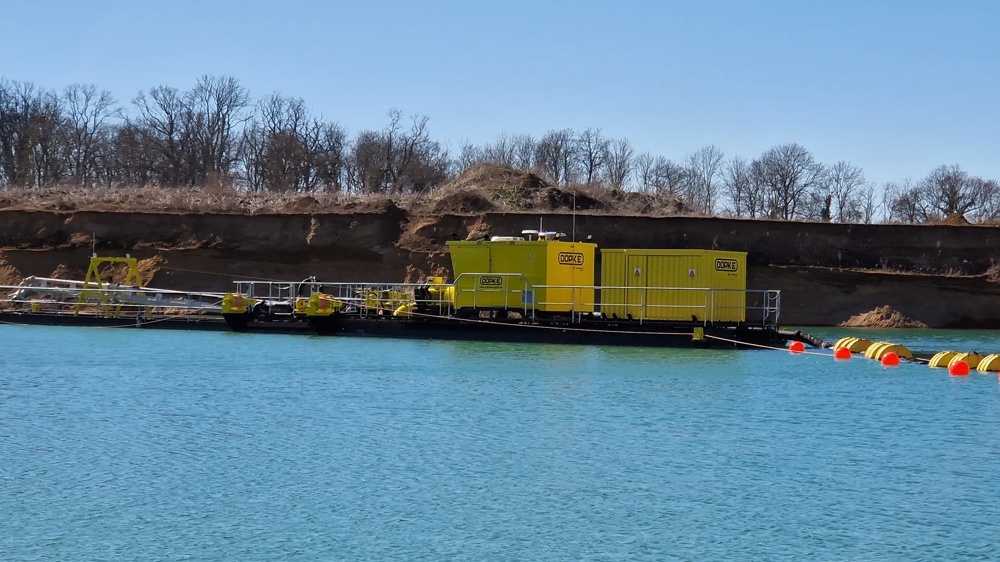 Gelbes Industriegebäude auf einem Floß im blauen Wasser, mit kahlen Bäumen im Hintergrund.