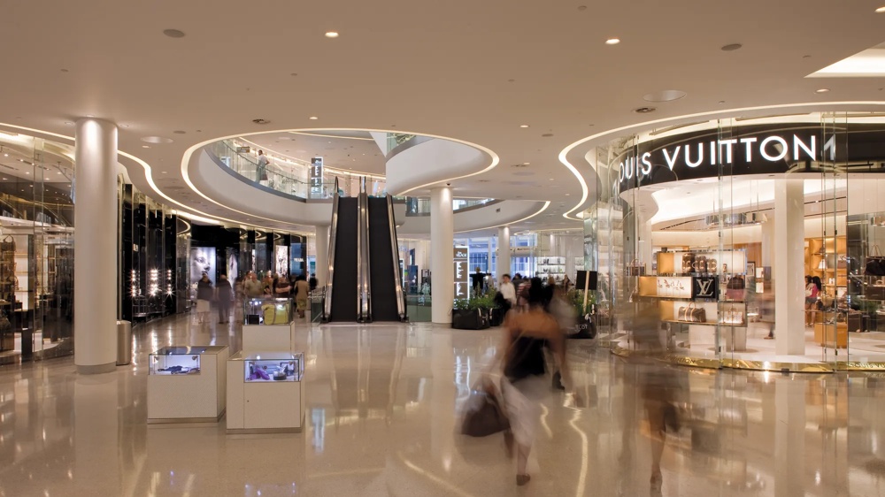 Ein modernes Einkaufszentrum mit glänzendem Boden, Rolltreppen und einem Louis Vuitton-Geschäft rechts.
