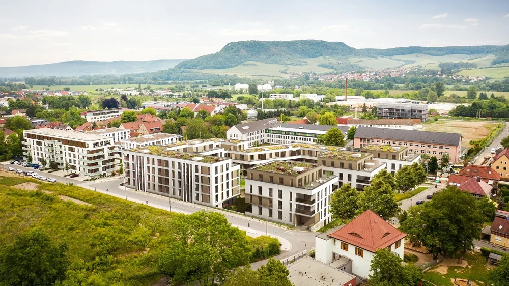Modern residential building with green roofs in front of a hilly landscape and a small village in the background.