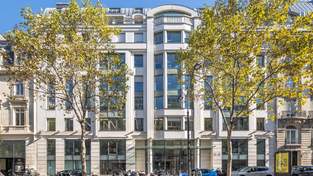 Weiße Fassade eines modernen Gebäudes, umgeben von grünen Bäumen unter blauem Himmel.