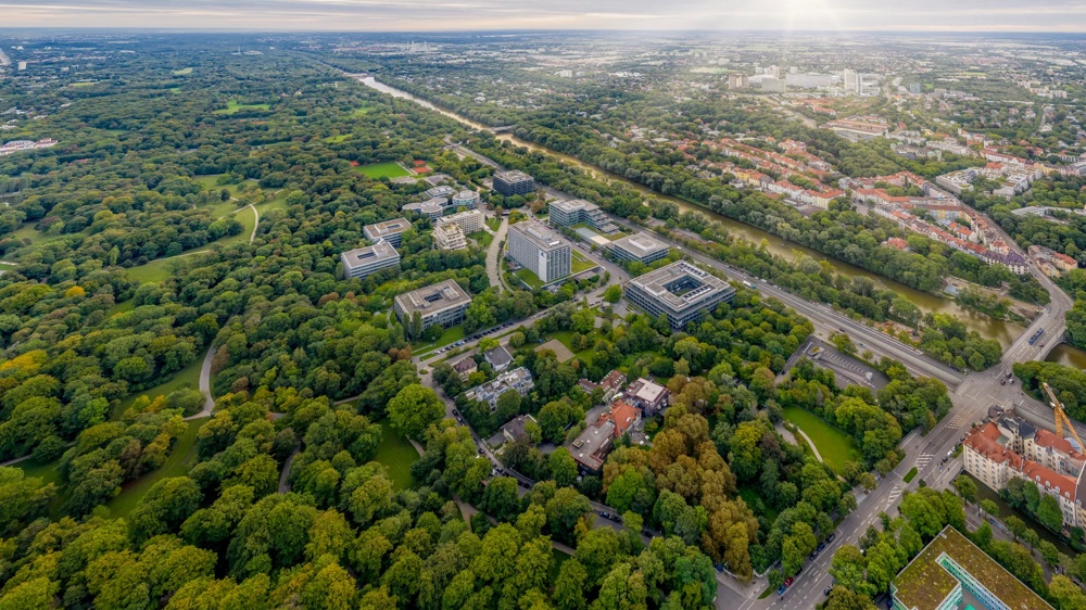 Luftaufnahme eines weitläufigen Parks mit vielen Bäumen, Gebäuden und einem Fluss unter bewölktem Himmel.