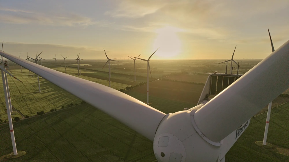 Nahaufnahme einer Windkraftanlage im Sonnenuntergang, umgeben von grünen Feldern und weiteren Windrädern.