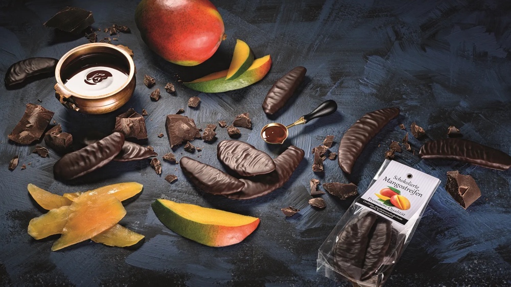 Schokolierte Mangostreifen und frische Mango auf dunklem Untergrund, umgeben von Schokoladenstücken.