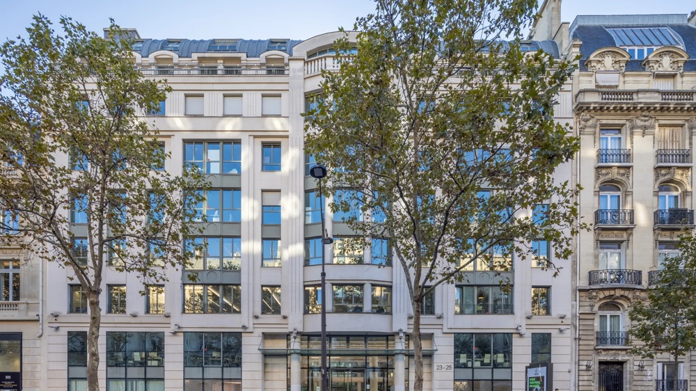 Moderne Fassade eines Bürogebäudes mit großen Fenstern, Bäumen und geparkten Motorrädern davor.