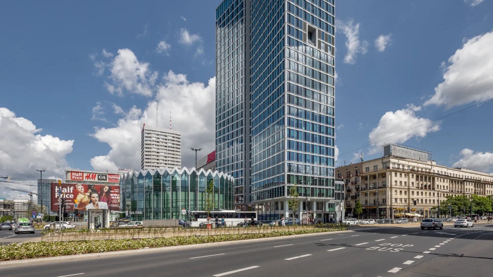 Hoher moderner Wolkenkratzer mit Glasfassade, umgeben von älteren Gebäuden und Werbetafeln unter blauem Himmel mit Wolken.