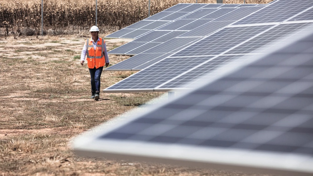 Eine Person in Warnweste und Helm geht neben einer Reihe von Solarpaneelen auf einem trockenen Feld.