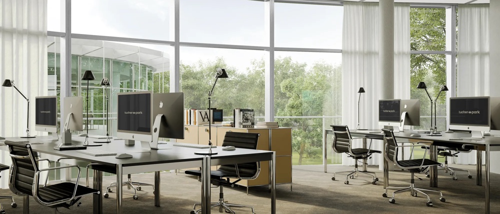 Modernes Büro mit großen Fenstern, mehreren Schreibtischen, Computern und Bürostühlen, umgeben von grüner Landschaft.