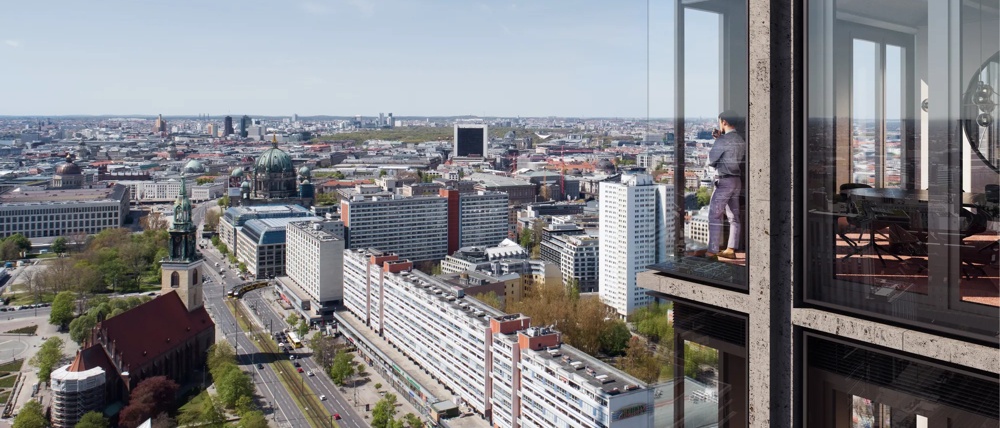 Ein Mann steht auf einem verglasten Balkon eines Hochhauses mit Blick auf eine Stadtlandschaft bei klarem Himmel.
