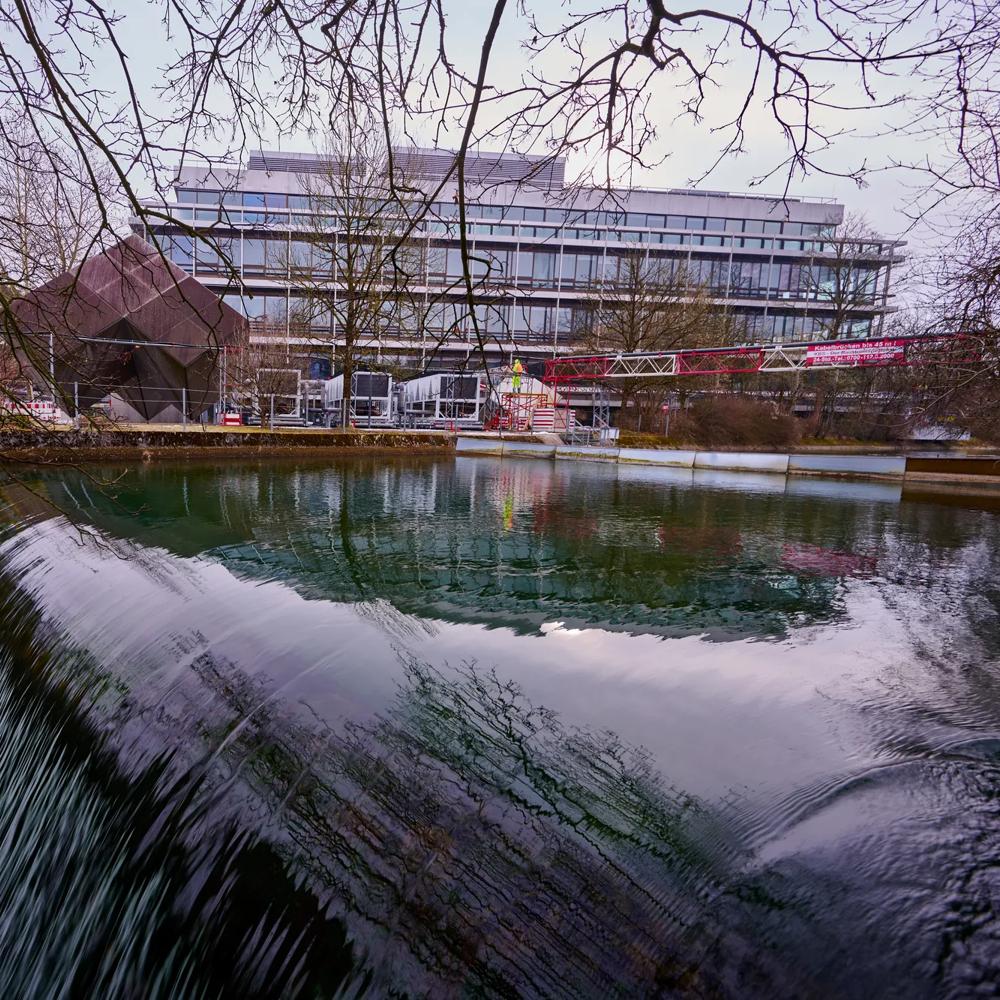 Wehrstufe an einem Fluss mit türkisfarbenem Wasser im Vordergrund. Im Hintergrund ein modernes Bürogebäude und eine markante dunkle Skulptur, eingerahmt von kahlen Winterbäumen.