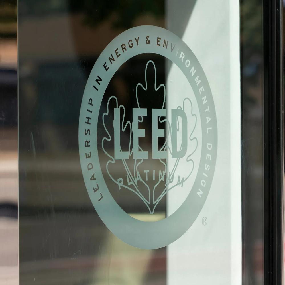 A glass door with a metal frame featuring a white round emblem with an oak leaf and the word “Leed,” representing real estate with high sustainability standards.