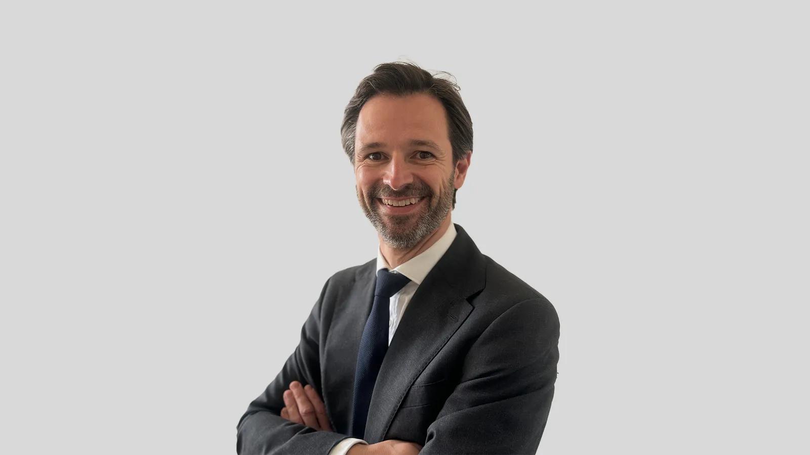 Ein lächelnder Mann im Anzug mit verschränkten Armen vor grauem Hintergrund.