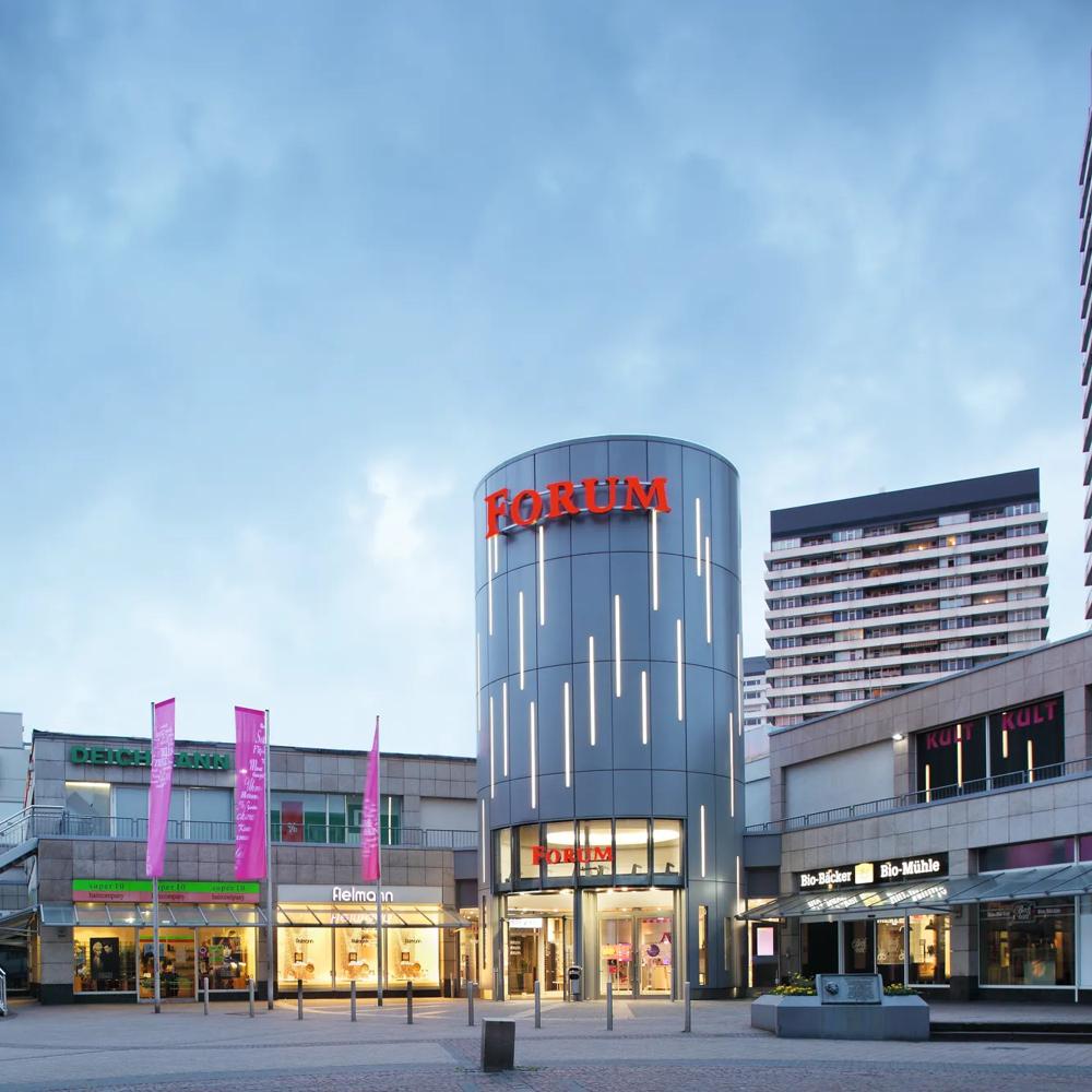 Einkaufszentrum mit grauem Turm, rotem "Forum" Schriftzug, umgeben von Geschäften und hohen Gebäuden.