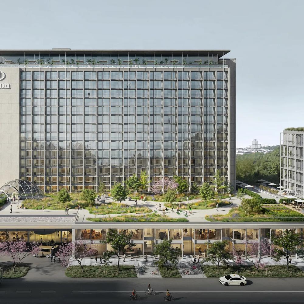 A large hotel building with a glass facade, green roof and surrounding trees, with a lively street in front.
