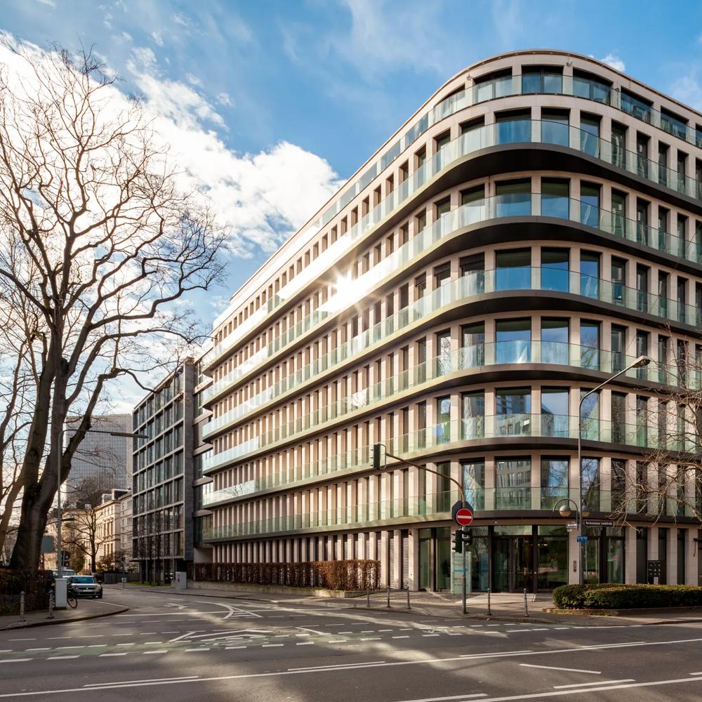 Modernes Bürogebäude mit Glasfassade an einer Straßenecke, blauer Himmel und kahle Bäume im Vordergrund.