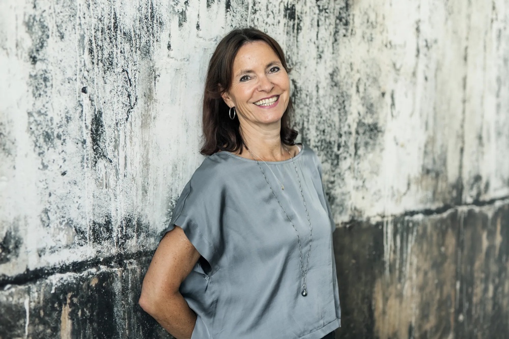 Eine lächelnde Frau in grauer Bluse lehnt an einer strukturierten, verwitterten Wand.