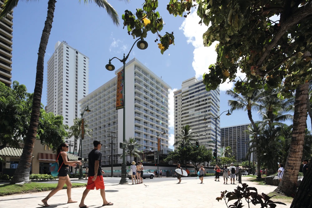 Straßenszene mit hohen Gebäuden, Palmen, Fußgängern und einem Surfer unter blauem Himmel.