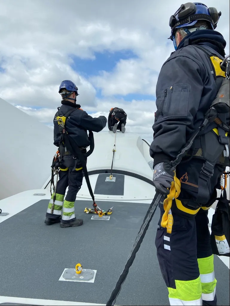 Zwei Arbeiter in Sicherheitsausrüstung stehen auf einer Windkraftanlage unter bewölktem Himmel.