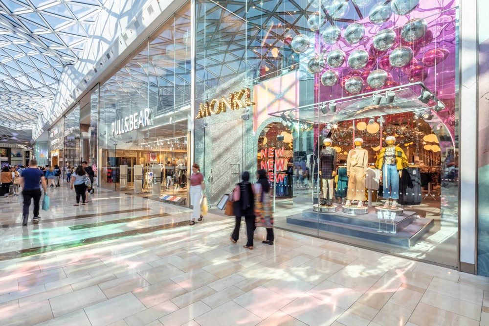 Einkaufszentrum mit gläsernem Dach, Geschäften und Schaufensterpuppen in bunter Beleuchtung, Menschen flanieren.