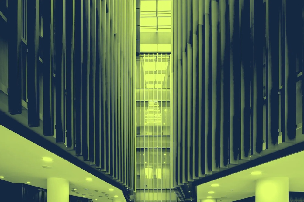 Moderner Innenraum mit hohen Lamellenwänden und Deckenlichtern, zentraler Glasaufzug im Hintergrund.