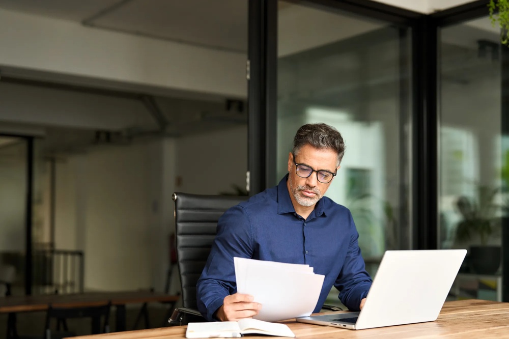 Ein Mann mit Brille liest Papiere am Schreibtisch mit Laptop in modernem Büro.