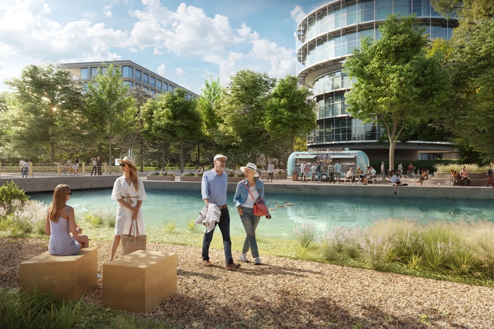 Menschen spazieren an einem Teich mit Enten, Bäumen und modernen Gebäuden im Hintergrund.