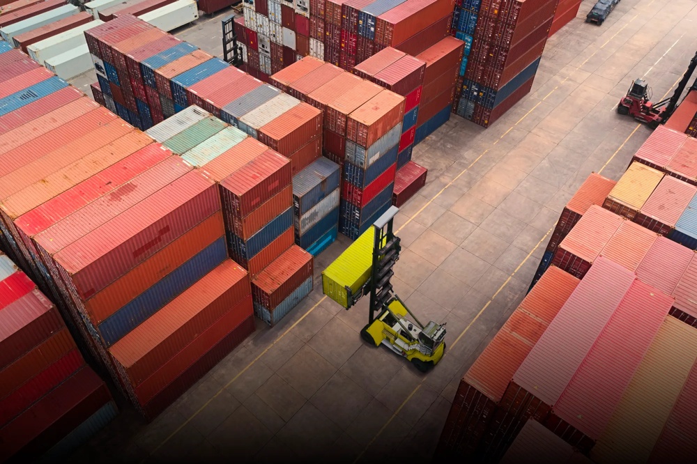 Ein gelber Gabelstapler hebt einen Container zwischen gestapelten bunten Containern in einem Hafenlager.