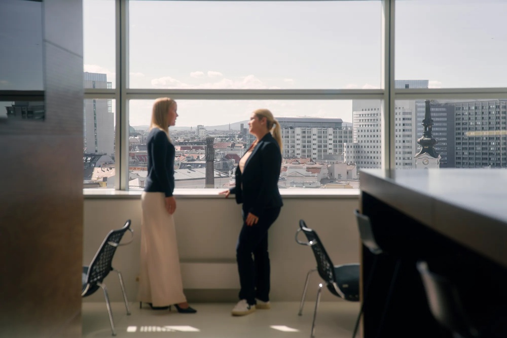 Zwei Personen stehen vor einem großen Fenster mit Stadtblick und unterhalten sich.