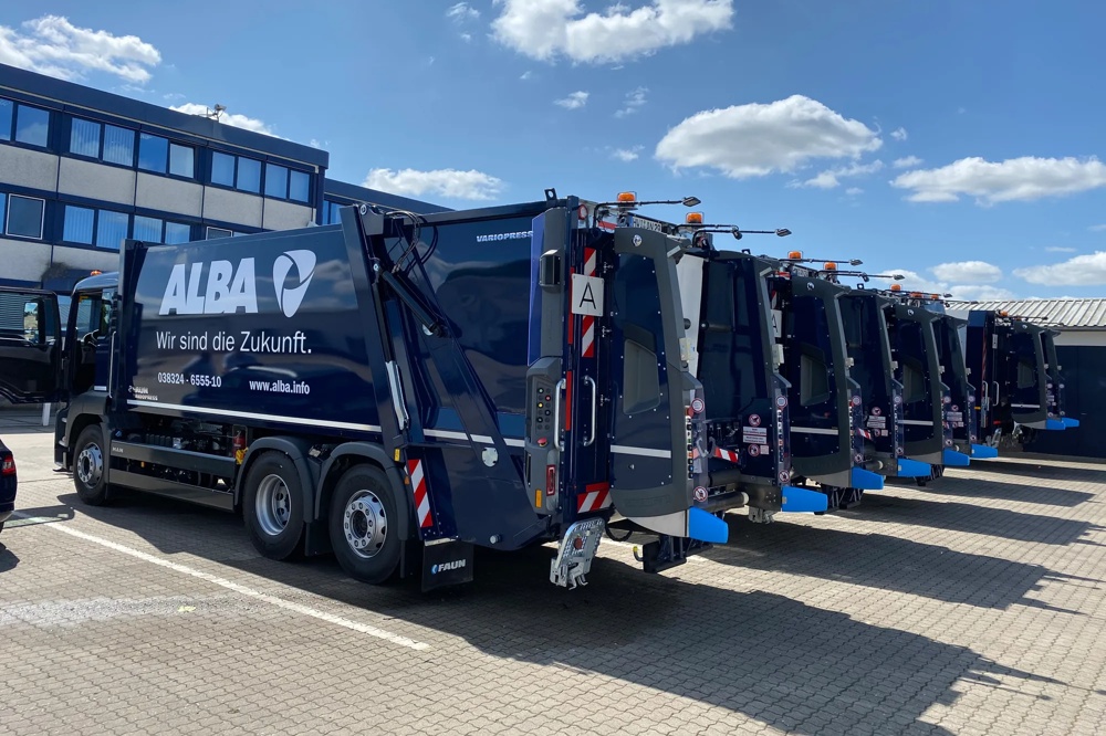 Eine Reihe von blauen Müllwagen auf einem gepflasterten Parkplatz bei sonnigem Wetter.