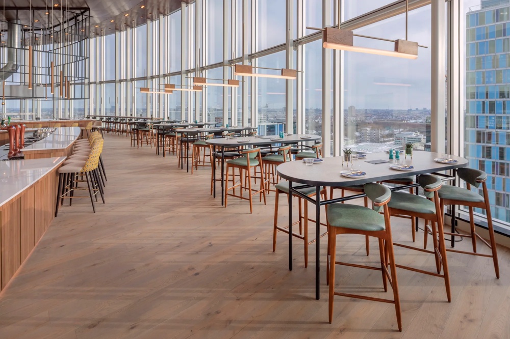 Ein modernes Restaurant mit hohen Fenstern, Holztischen und grünen Stühlen, bietet einen weiten Stadtblick.