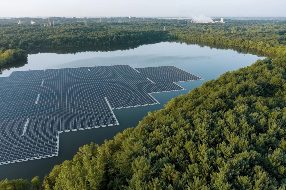 Schwimmende Solaranlage auf einem See, umgeben von dichtem Wald, mit Fabrik im Hintergrund.
