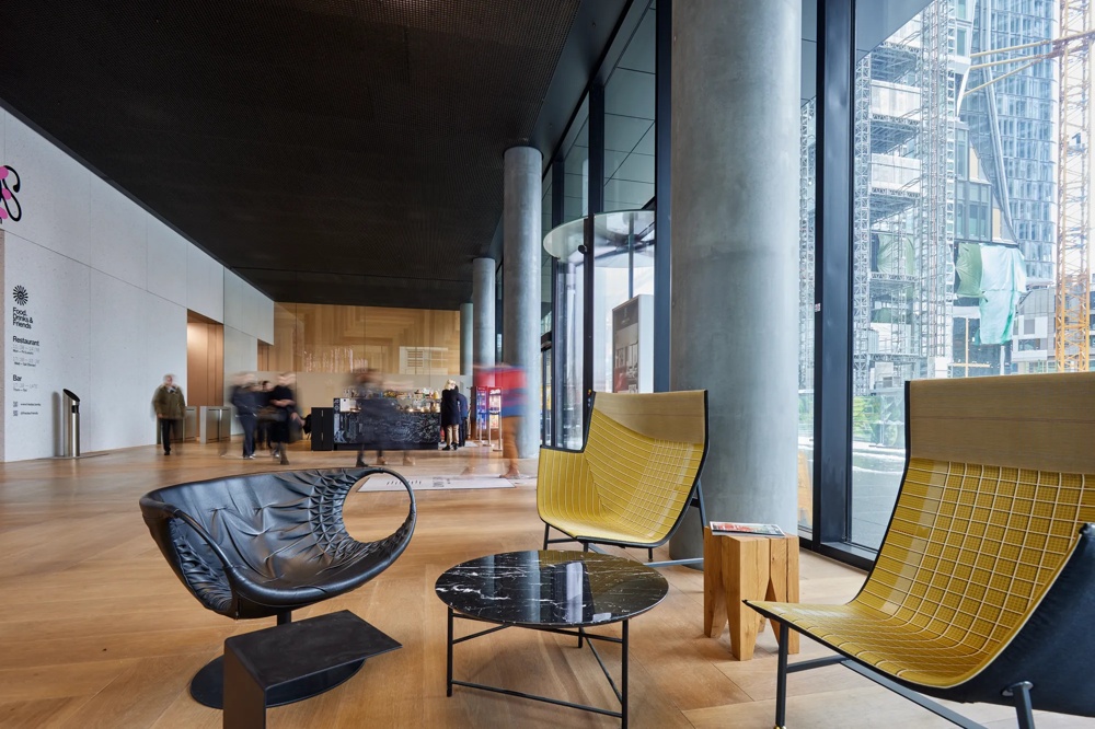 Moderne Lobby mit gelben Sesseln, rundem Marmortisch und hohen Glasfenstern, Menschen im Hintergrund, Holzfußboden.